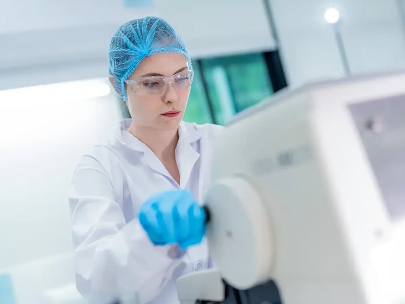 A focused laboratory technician in a white coat, blue gloves, and a hair net operates scientific equipment in a bright, sterile lab environment