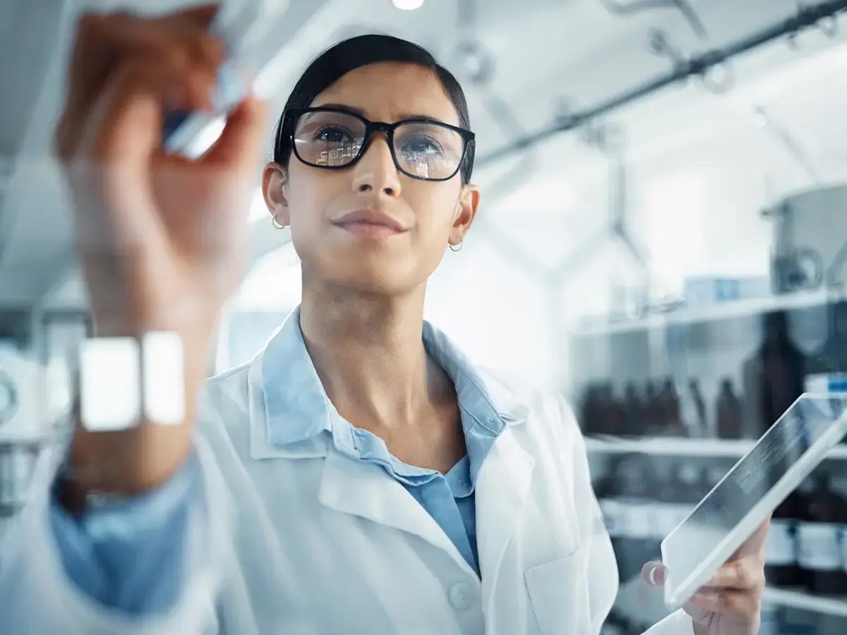 A focused scientist in a lab coat and glasses writes on a transparent board while holding a tablet