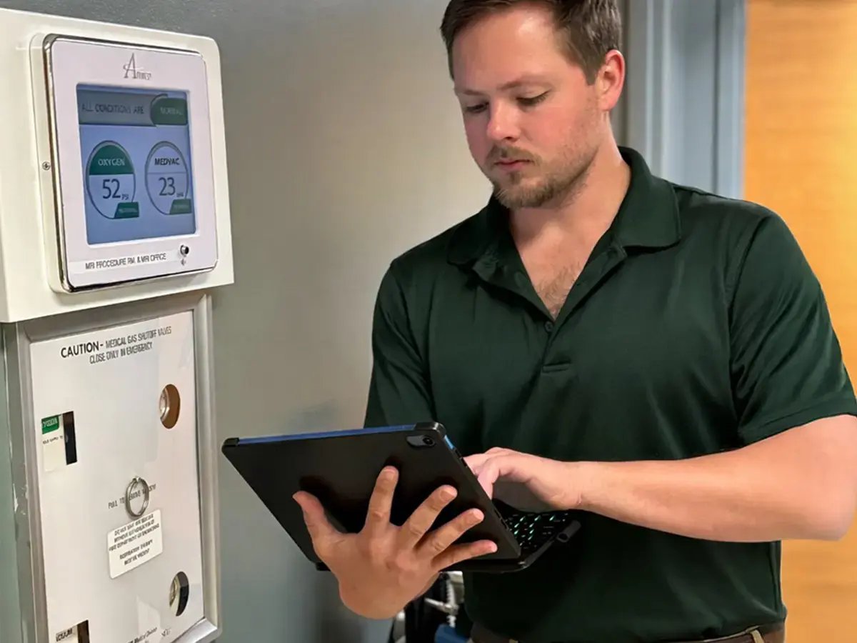 A man in a green polo uses a tablet next to a medical control panel displaying oxygen and medical air levels
