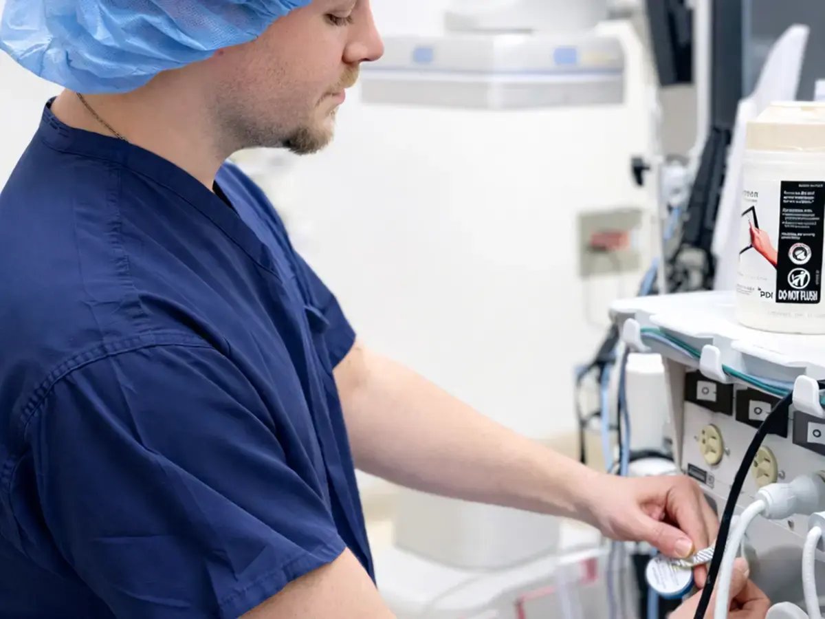 A medical professional in blue scrubs and a hair net adjusts equipment in a hospital setting