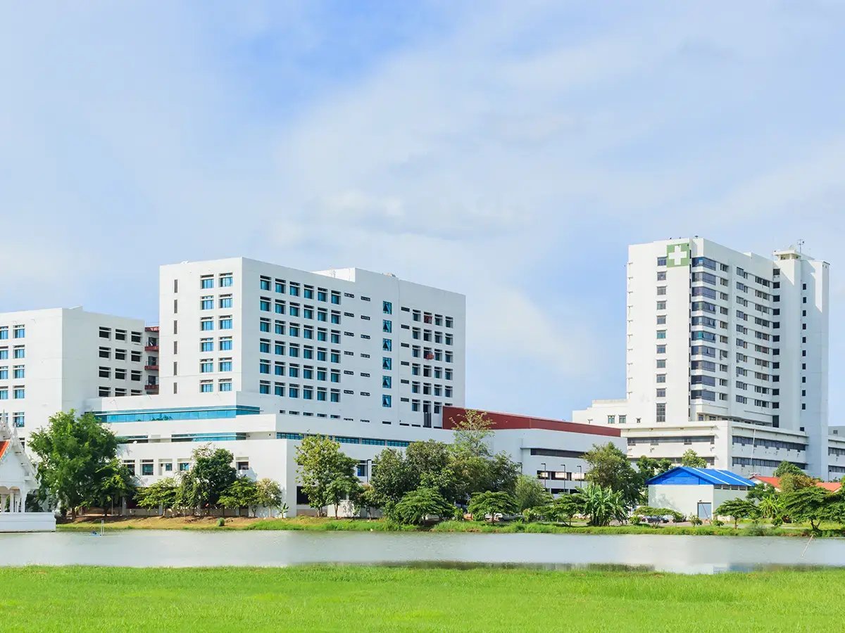 A modern, large hospital with multiple white buildings surrounded by greenery and a river under a clear blue sky