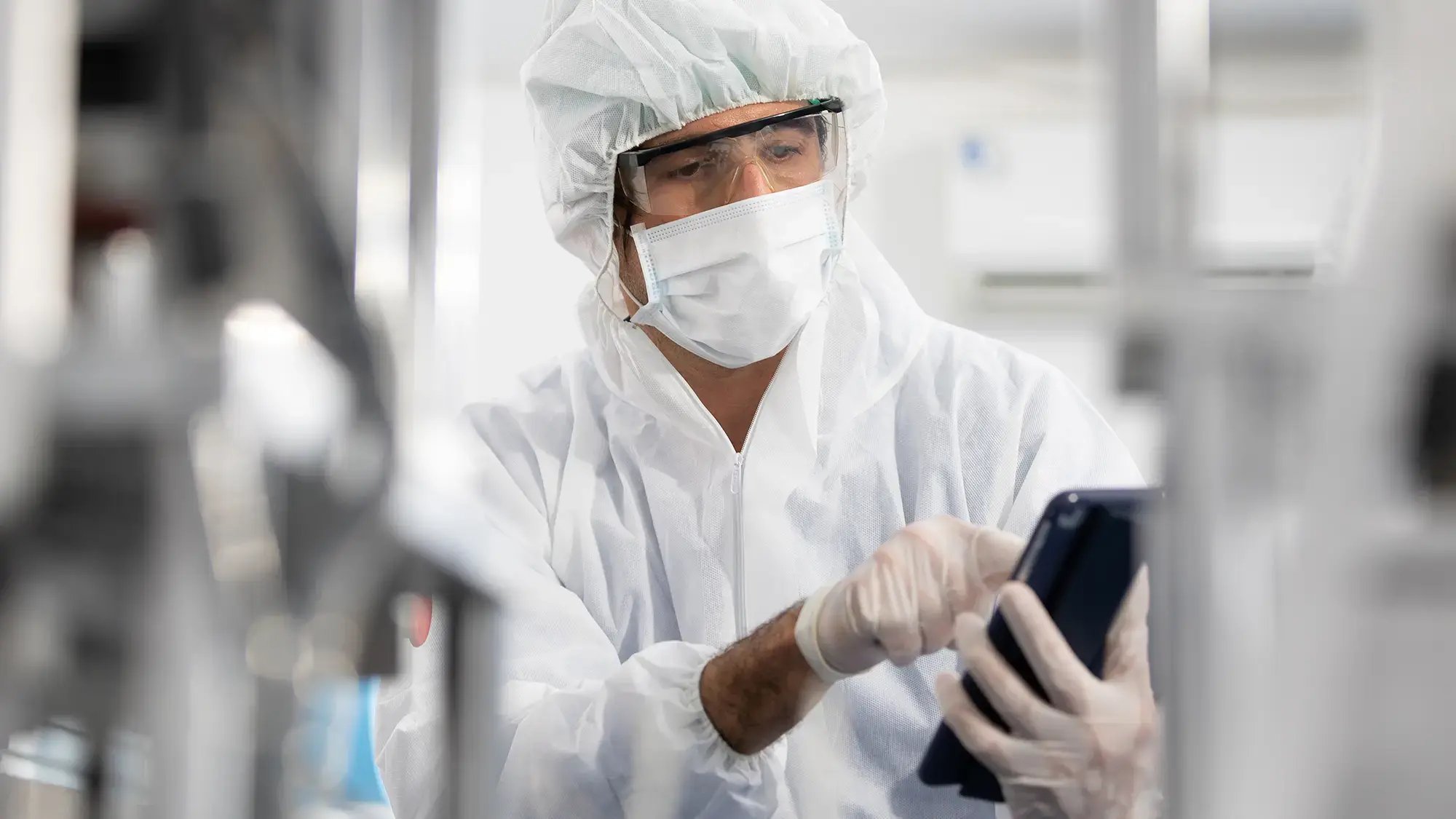 A person in a clean room suit, mask, and goggles operates a device in a lab setting