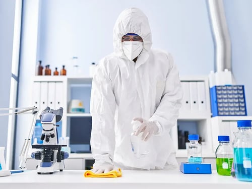 A person in full protective gear cleans a laboratory table with a spray bottle and cloth