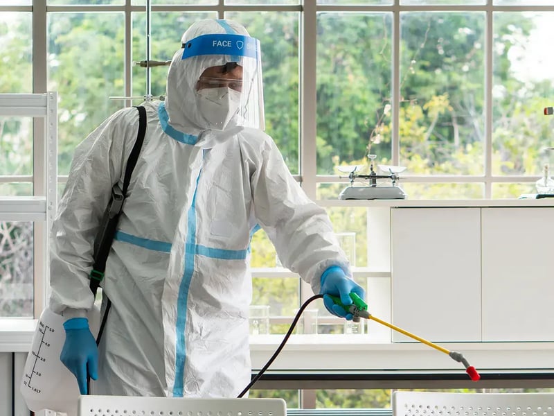 A person in full protective gear, including a face shield and gloves, is disinfecting a bright room with large windows, holding a spray nozzle