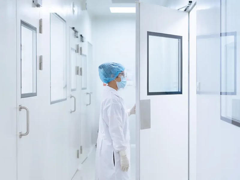 A person in medical attire, including a lab coat, mask, and hairnet, opens a door in a sterile lab, suggesting a clean, controlled setting