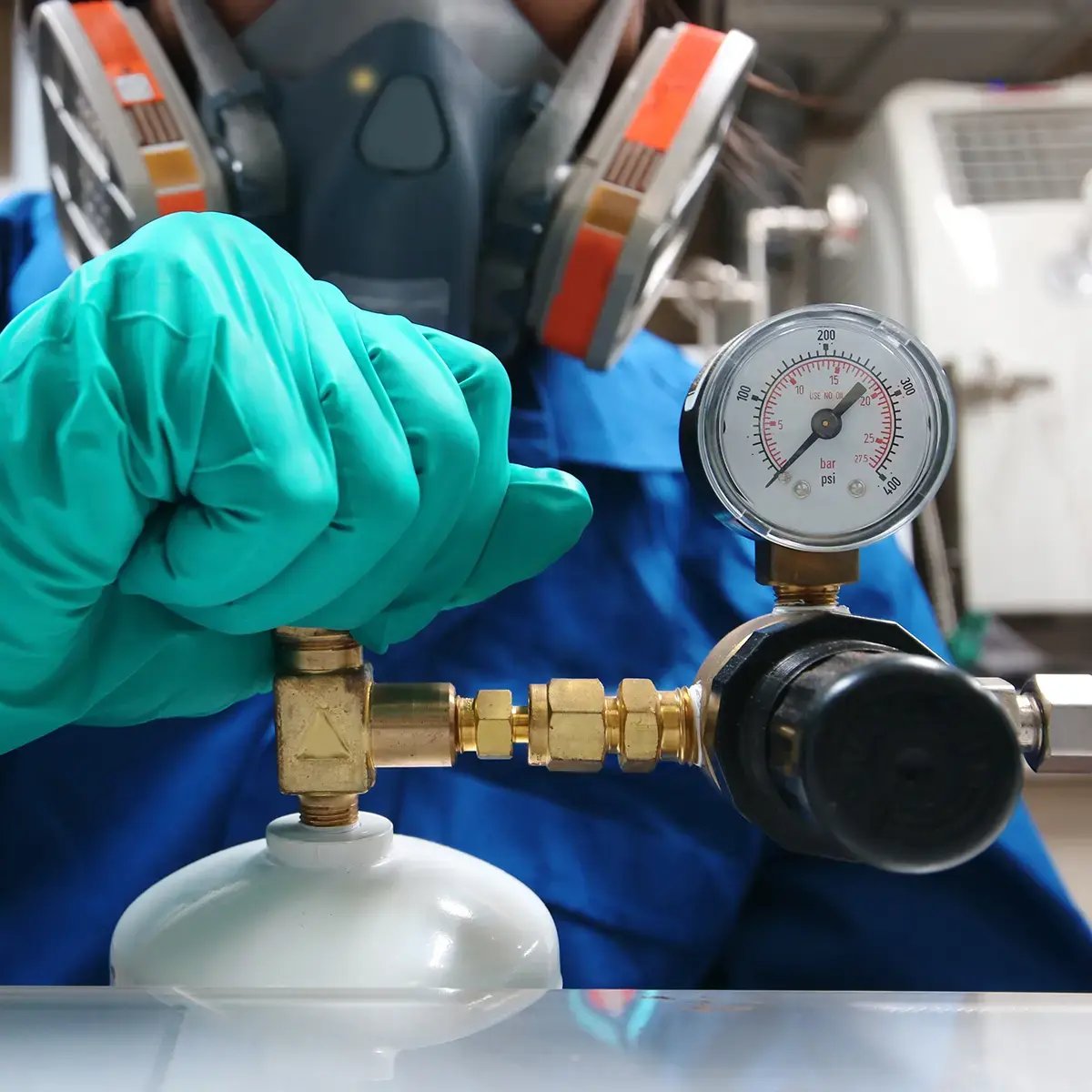 A person wearing a protective mask and green gloves adjusts a gas pressure regulator with a dial gauge in a lab, conveying caution and precision