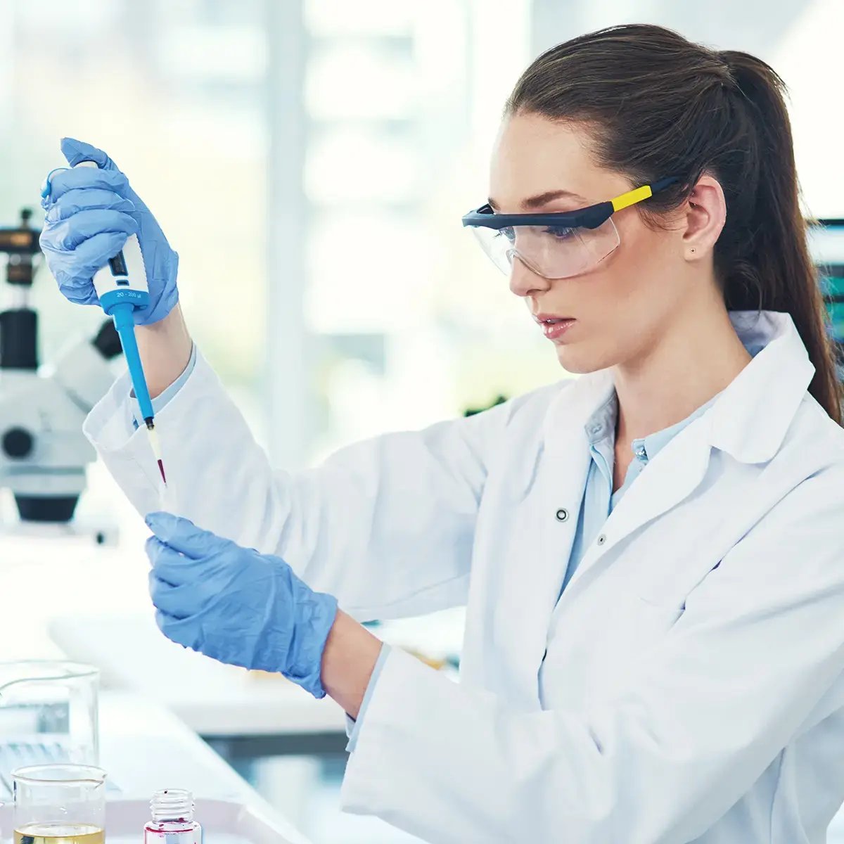 A scientist in a lab coat and safety goggles concentrates while using a pipette