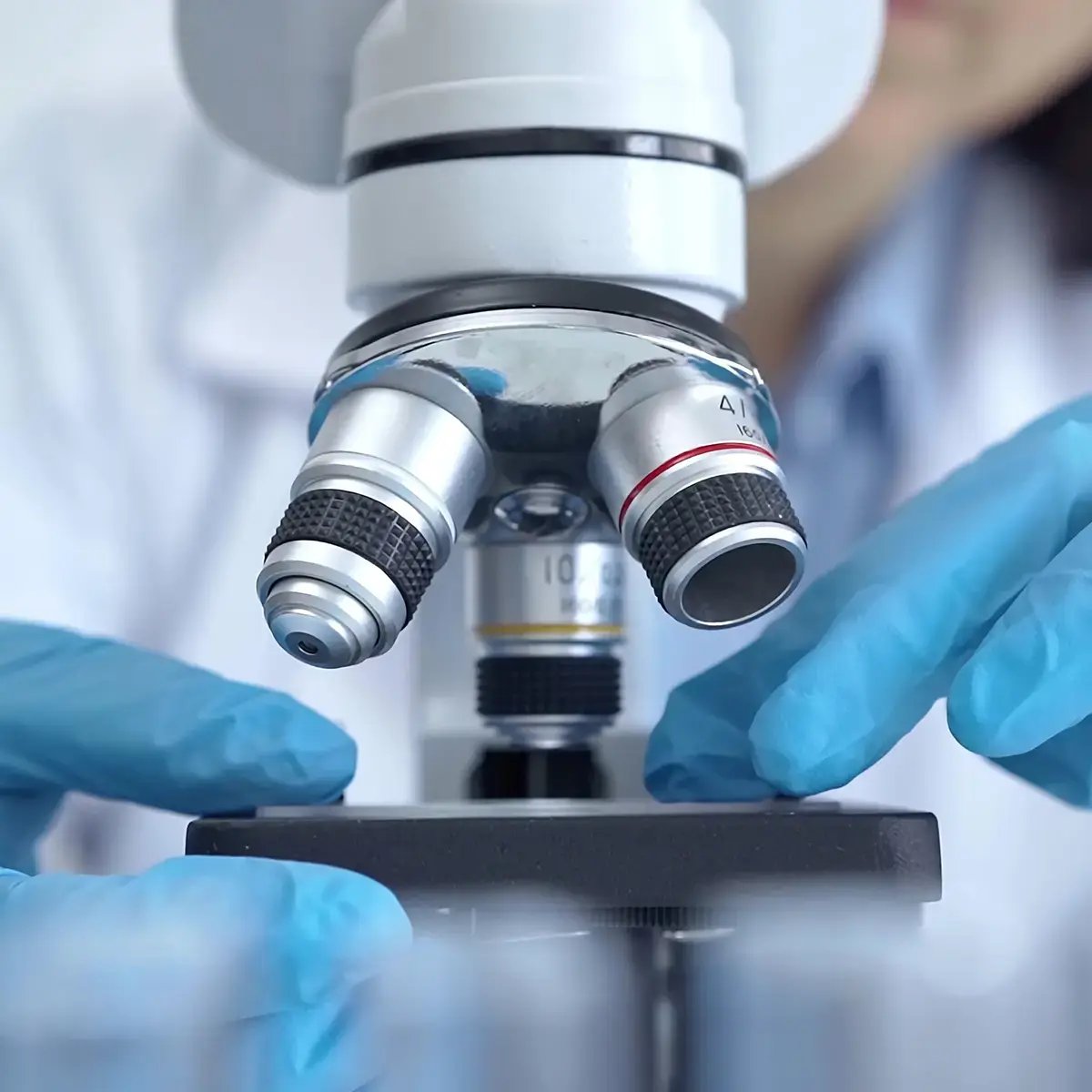 A scientist in blue gloves adjusts a microscope, focusing on intricate lenses