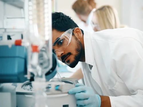 A scientist wearing goggles and gloves closely examines laboratory equipment