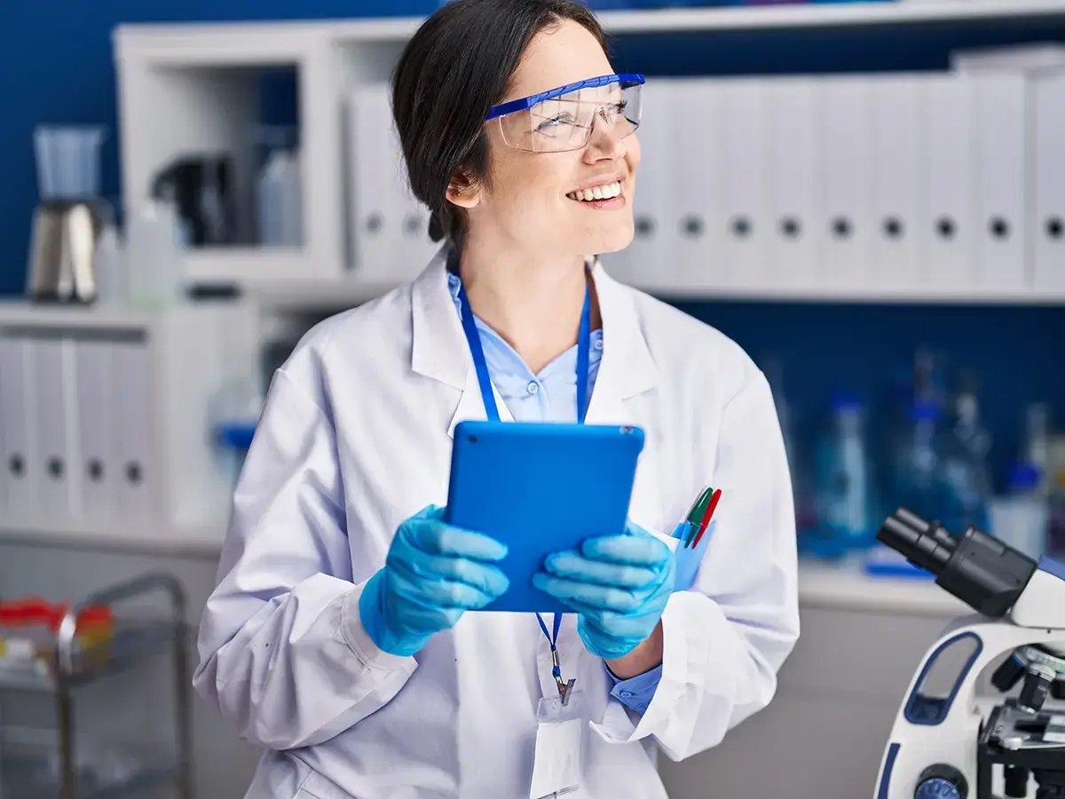 A smiling scientist in a lab coat, gloves, and safety goggles holds a blue tablet