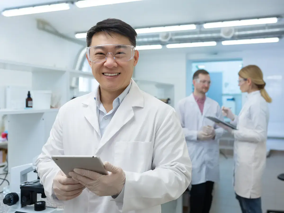 A smiling scientist in safety goggles holds a tablet in a lab