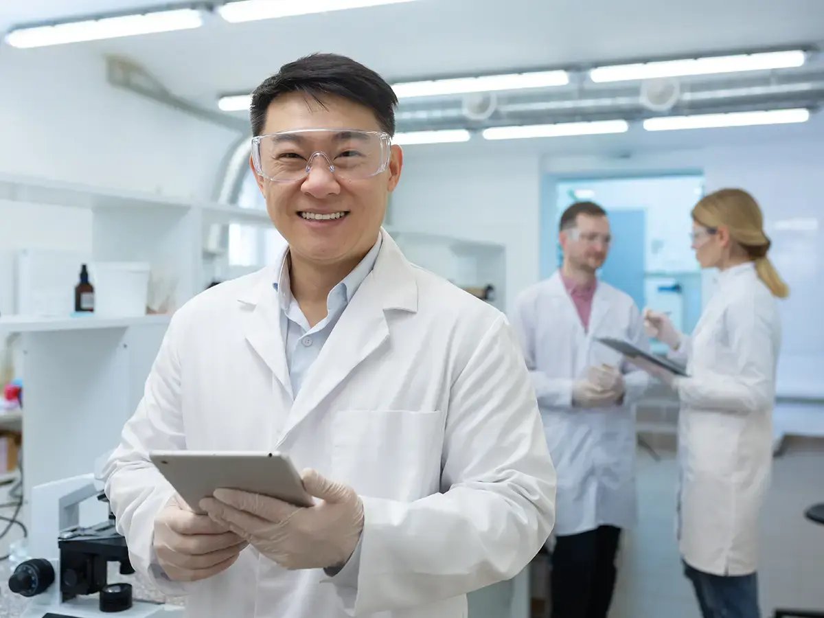 A smiling scientist in safety goggles holds a tablet in a lab