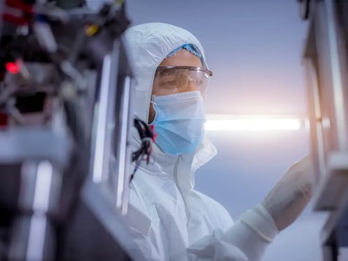 A technician in a clean room suit, mask, and goggles operates equipment in a sterile lab