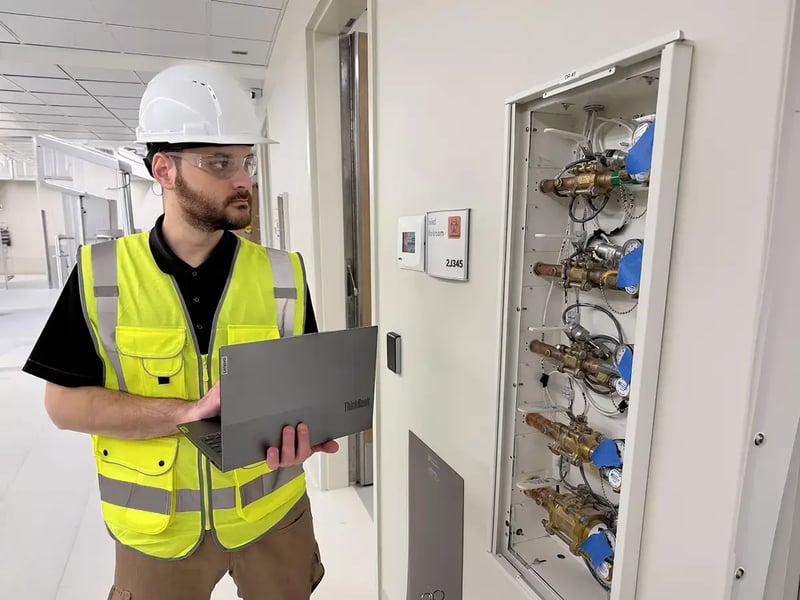 A worker in a white hard hat and yellow safety vest inspects a meter panel while holding a laptop