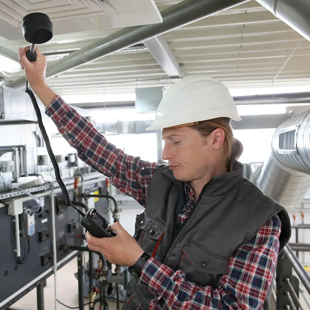 A worker wearing a hard hat and plaid shirt holds a measuring device up to an air vent in an industrial setting