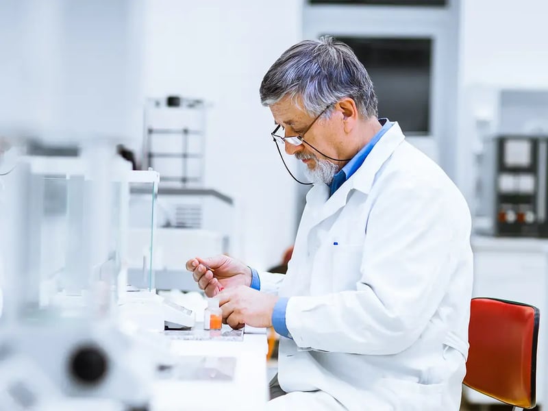 Elderly male scientist in a white lab coat focuses intently on an experiment in a bright, modern laboratory, conveying concentration and expertise