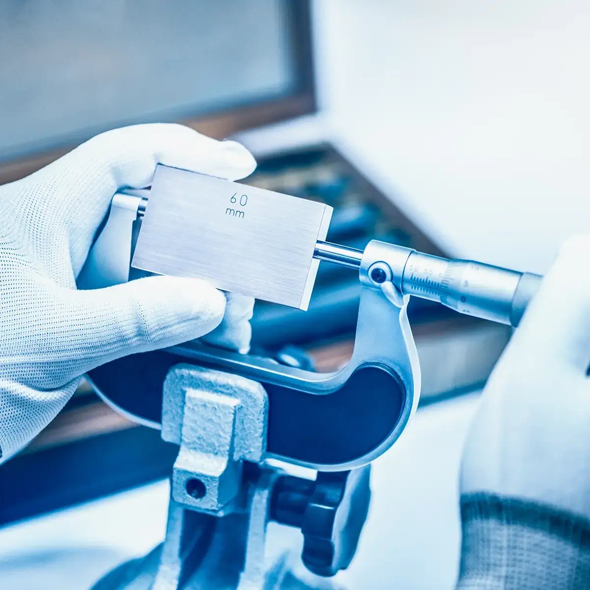 Gloved hands hold a micrometer measuring a 60 mm metal block, conveying precision and focus in an engineering or laboratory setting