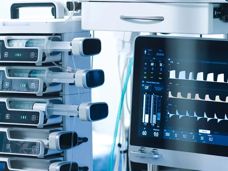Medical equipment in a hospital includes an infusion pump with syringes on the left and a display screen showing vital signs and waveforms on the right