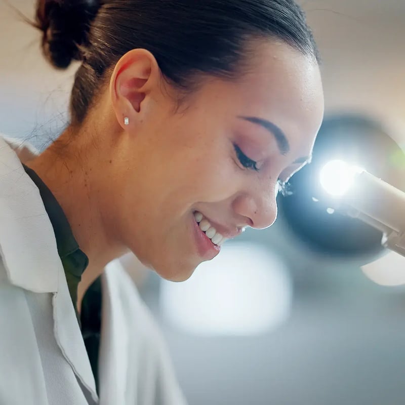 Smiling woman in a lab coat examines a sample under a microscope, conveying focus and positivity in a bright, scientific setting