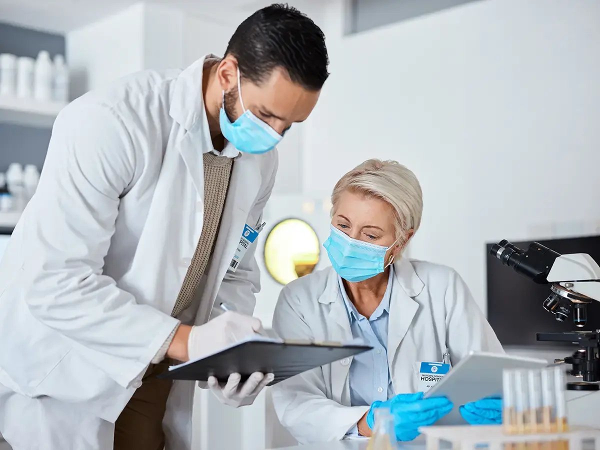 Two healthcare professionals in lab coats and masks discuss data in a laboratory