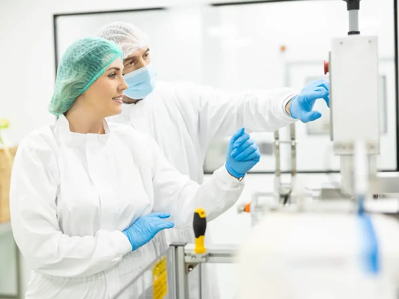 Two laboratory technicians in white protective suits and blue gloves operate machinery