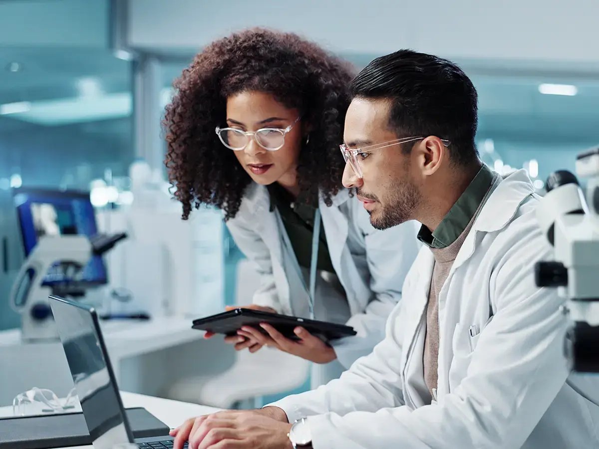 Two scientists in lab coats, one holding a tablet, examine data on a laptop in a modern laboratory