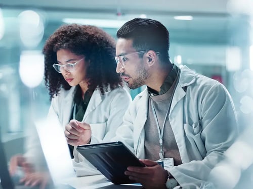 Two scientists in white lab coats focus on data on a tablet in a bright laboratory