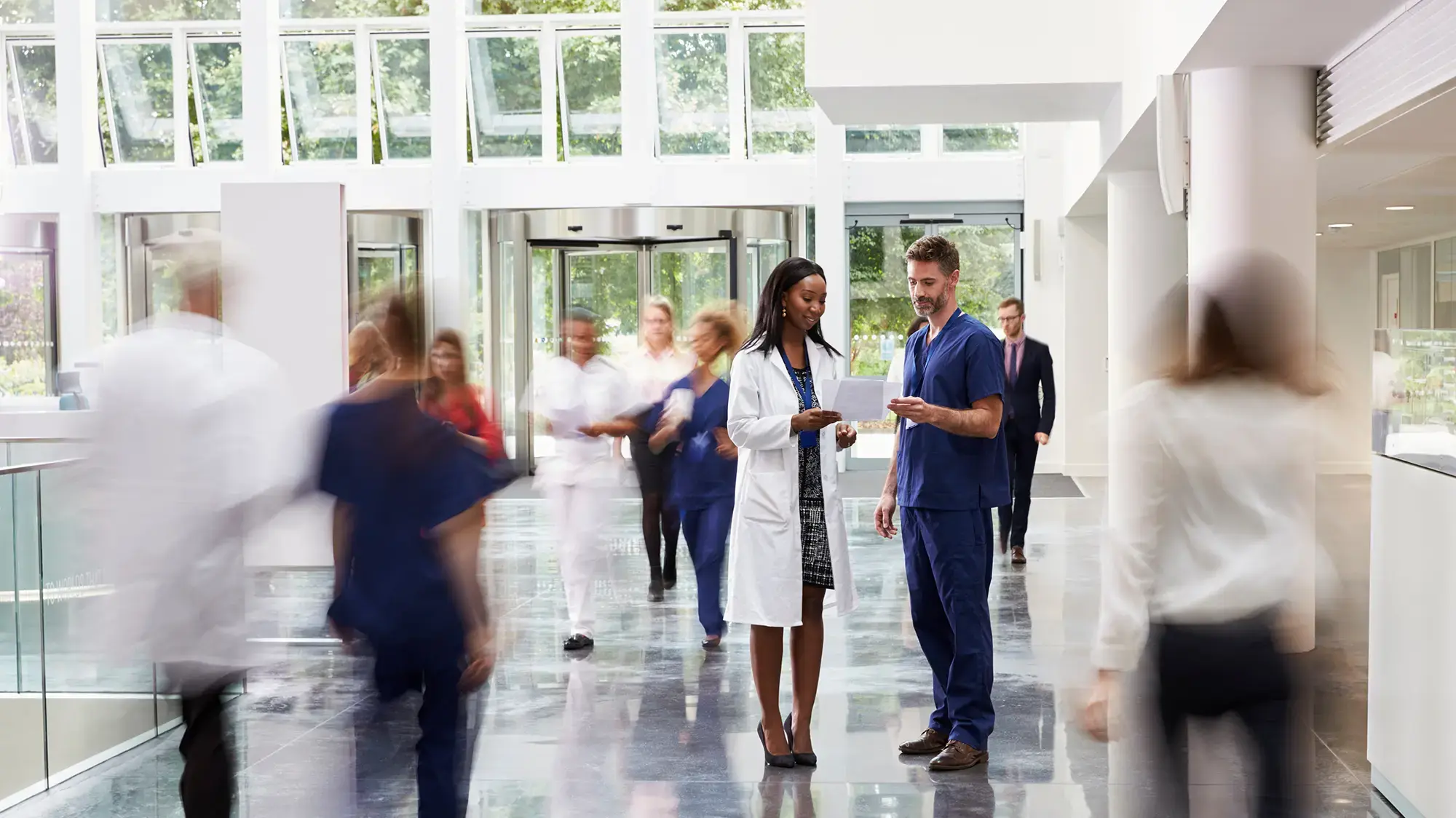 Hospital lobby with doctors and nurses in motion; a female doctor and a male nurse discuss a document in focus